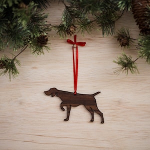 May include: A wooden Christmas ornament shaped like a German Shorthaired Pointer dog. The ornament is brown and has a red ribbon attached to it.