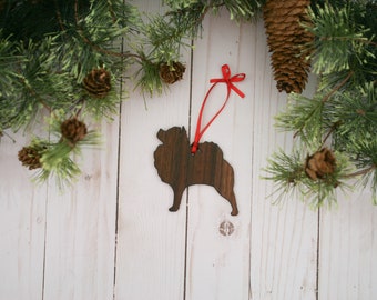 May include: Wooden Christmas ornament shaped like a Pomeranian dog, painted brown, with a red ribbon attached. The ornament is hanging from a pine branch with pine cones.