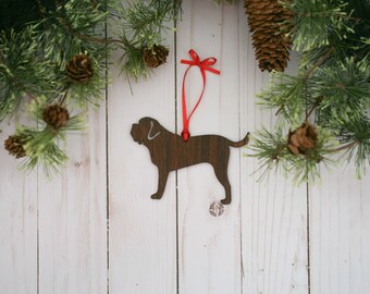 May include: A wooden Christmas ornament shaped like a brown dog with a red ribbon attached. The ornament is hanging from a pine branch with pine cones.