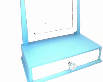 May include: A light blue vanity mirror with a small drawer and a white knob. The mirror is attached to a stand with two vertical supports.