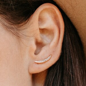May include: A close-up of a person's ear with a small gold crescent moon earring with small clear stones.