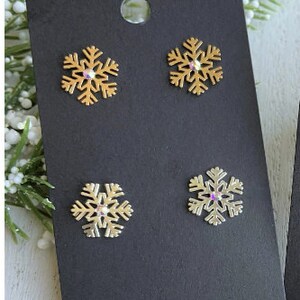 May include: A black card with four snowflake earrings. Two of the earrings are gold with clear rhinestones and two are silver with clear rhinestones.