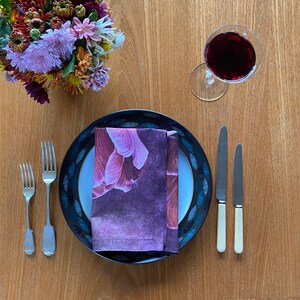 May include: A black and white patterned dinner plate with a purple floral napkin on top. Two silver forks and two silver knives are placed on the table next to the plate. A glass of red wine is on the table to the right of the plate.