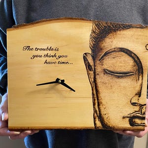 May include: Wooden clock with a natural edge, featuring a burned-in image of Buddha's face on the right side. The clock face has the quote "The trouble is you think you have time..." and black clock hands.