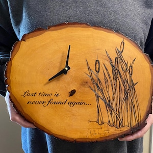 May include: A round wooden clock with a natural edge. The clock face is light brown with black clock hands. The clock features an etched design of cattails and the quote, "Lost time is never found again..."