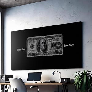 May include: A modern office space with a large black artwork featuring a grayscale $100 bill with the text "Money first. Love later." A desk with a laptop, speakers, and a desk lamp is in front of the artwork. Potted plants and a gray office chair are also visible.