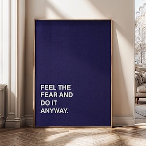 May include: A framed print with a dark blue backdrop and the white text "FEEL THE FEAR AND DO IT ANYWAY." The artwork is in a gold-coloured frame and is displayed on a wooden floor.