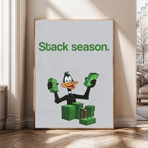 Op de afbeelding: Een ingelijste print met Daffy Duck die stapels geld vasthoudt. De zin "Stack season" is in het groen boven het stripfiguur gedrukt. De achtergrond is lichtgrijs.