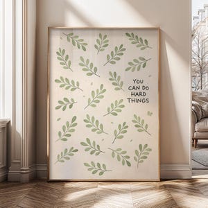 Może przedstawiać: Oprawiony wydruk z beżowym tłem ozdobionym ilustracjami zielonych liści. Czarny tekst brzmi "YOU CAN DO HARD THINGS". Rama ma jasnobrązowy kolor.