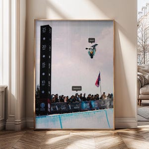 Op de afbeelding: Ingelijste kunstdruk met een snowboarder in de lucht boven een menigte. Een zwarte meetpaal geeft hoogtes tot 7,6 meter aan. De woorden "dreamers" en "doers" zijn aanwezig. Een rood-paarse vlag wappert op de achtergrond.