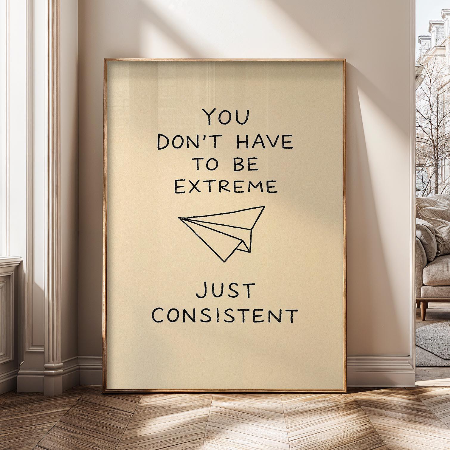 May include: A framed print with a tan background and gold frame. The black text reads "YOU DON'T HAVE TO BE EXTREME" above a paper airplane illustration, and "JUST CONSISTENT" below. The print is displayed in a room.