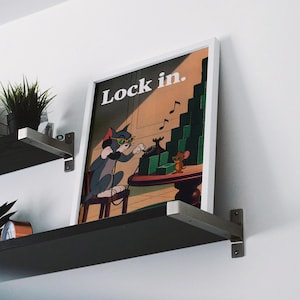 May include: Framed art print featuring Tom from Tom and Jerry wearing sunglasses and headphones, dialing a rotary phone. The print has the words "Lock in." and a stack of money. The print is displayed on a shelf with a plant.