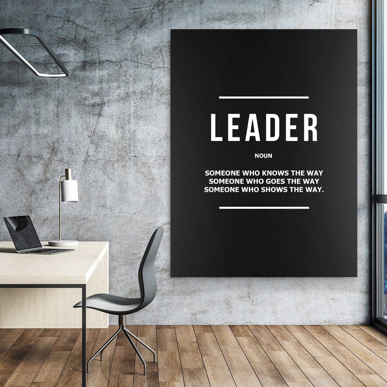May include: A black and white poster with the word 'LEADER' in large white letters. Below the word is the word 'NOUN' in smaller white letters. Below the word 'NOUN' is the text 'SOMEONE WHO KNOWS THE WAY', 'SOMEONE WHO GOES THE WAY', and 'SOMEONE WHO SHOWS THE WAY.' The poster is hanging on a gray concrete wall.