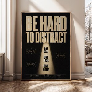 Op de afbeelding: Een rechthoekige wandposter met een zwarte achtergrond en gouden tekst. Bovenaan staat BE HARD TO DISTRACT. Daaronder belicht een spotlight-effect de woorden ONE GOAL, ONE TASK, ONE HOUR. Doorgestreepte woorden zijn NOTIFICATIONS, OPINIONS en URGENCY.