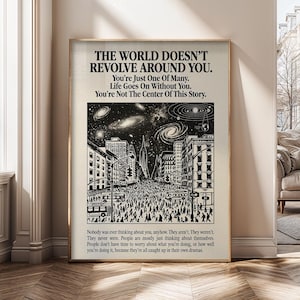 May include: A framed poster with a beige background and black text. The text reads "THE WORLD DOESN'T REVOLVE AROUND YOU." Below is a black and white illustration of a city street filled with people. Additional text is at the bottom.