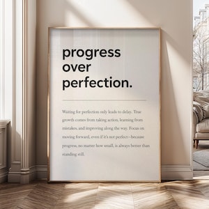 May include: A framed print with the words "progress over perfection" in a bold, black font. Below the title is a paragraph explaining the concept. The frame is a light brown color.