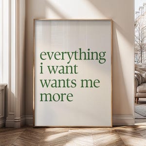 May include: A framed art print featuring the phrase "everything i want wants me more" in green serif font. The print has a cream background and a gold frame. The artwork is displayed in a room with natural light, creating a minimalist aesthetic.