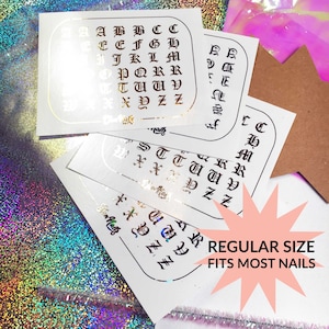 May include: Four sheets of white paper with gold letters printed on them. The letters are in a gothic font and spell out the alphabet. The sheets are stacked on top of each other. The text "REGULAR SIZE FITS MOST NAILS" is printed on a pink starburst.
