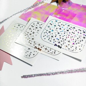 May include: Three sheets of white sticker paper with a pattern of stars in different sizes. The sheets are labeled "Div-Nails" in a gold, pink, and blue holographic foil.