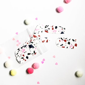 May include: A clear plastic bag filled with white, black, pink, and red terrazzo patterned nail wraps.  Two additional nail wraps are outside of the bag on a white background.