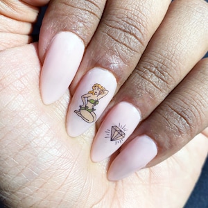 May include: Close-up of a hand with four nails painted a light pink color. Each nail has a different design. One nail has a diamond design, one has a pin-up girl design, and two have a solid pink color.