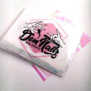 May include: A white envelope with a pink hexagon containing a black and white illustration of hands holding a nail file. The text "Dem Nails" is written in a black cursive font with the words "Nail Decals" below it.