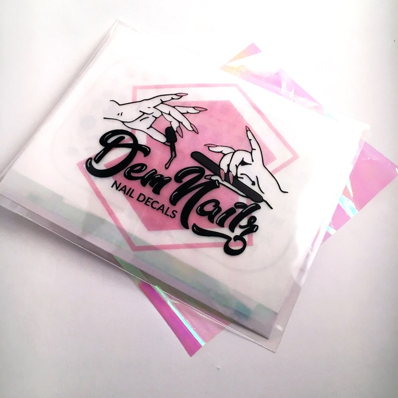 May include: A white envelope with a pink hexagon containing a black and white illustration of hands holding a nail file. The text "Dem Nails" is written in a black cursive font with the words "Nail Decals" below it.
