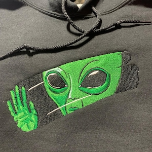 May include: Black hoodie with a green alien face embroidered on the front. The alien has large black eyes and is set against a black rectangular background. The hoodie has a drawstring hood.