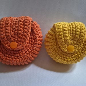 May include: Two handmade crochet coin purses. One is orange, the other is yellow. Each has a rounded shape with a flap and a button closure. The purses are made with a thick yarn, giving them a textured appearance.