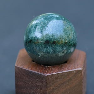 May include: A polished, spherical green stone with mottled dark inclusions and a horizontal band of small black spots, resting securely in a hexagonal, dark brown wooden stand with a circular cutout at the top center.