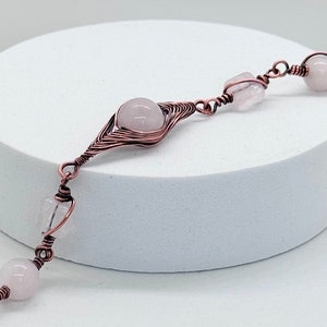 May include: A copper wire wrapped bracelet with pink rose quartz stones. The bracelet features a larger rose quartz stone in the centre, surrounded by smaller stones.