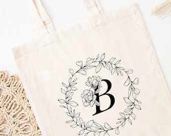 May include: A white canvas tote bag with black floral wreath and the letter "B" in the center.