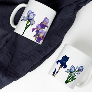 May include: Two white ceramic mugs with floral designs. One mug features purple and blue irises, while the other mug features blue and white irises.