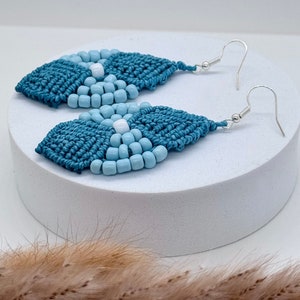 May include: Teal and light blue beaded earrings with silver hooks. The earrings are in the shape of a teardrop and have a geometric pattern.
