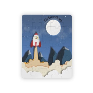Peut inclure: Un puzzle bleu et blanc représentant une fusée qui décolle dans le ciel nocturne avec une pleine lune et des étoiles. Le puzzle est intitulé "Sebastian".