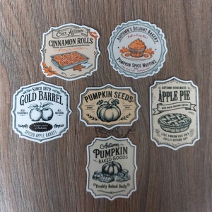 May include: Six vintage-style stickers with autumn-themed designs. The stickers feature illustrations of cinnamon rolls, pumpkin spice muffins, pumpkin seeds, apple pie, and pumpkin baked goods. The stickers have a rustic, hand-drawn aesthetic and feature text such as "Cozy Autumn", "Pumpkin Seeds", and "Autumn Homemade Apple Pie".