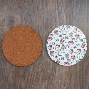 Op de afbeelding: Twee ronde onderzetters. Eén is gemaakt van kurk, de andere is wit met een herhalend patroon van koffiekopjes, boeken, strikken en bloemen in roze, rood en zwart. De onderzetters liggen op een houten oppervlak.