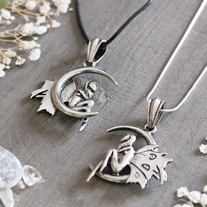 May include: Two silver necklaces with crescent moon pendants featuring fairies. One fairy is sitting on the moon with wings spread, the other is sitting on the moon with wings folded.