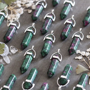 Peut inclure: Plusieurs pendentifs en pierres précieuses avec des capuchons en métal argenté. Les pendentifs sont un mélange de vert et de rouge, de forme hexagonale. Le fond est une surface grise avec de petites feuilles et des cristaux.