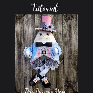 Puede incluir: Una muñeca blanca con forma de huevo que lleva un atuendo azul y rosa con una corbata de lazo negra y un sombrero a rayas. La muñeca está de pie sobre una superficie de madera. El texto "This Precious Mess" es visible en la parte inferior de la imagen.