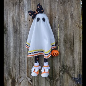 Op de afbeelding: Een witte spookpop met zwarte en oranje candy corn-bies, met een zwart-witte strik en oranje-witte candy corn-sokken. De pop houdt een kleine oranje pompoen vast.