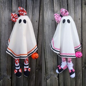 Op de afbeelding: Twee witte spookpoppen met zwarte ogen en zwart-wit gestreepte benen. E&eacute;n spookpop heeft een rode strik met een wit en oranje patroon en oranje-zwart gestreepte rand. De andere spookpop heeft een roze strik met een wit en roze patroon en roze-zwart gestreepte rand. Beide poppen houden oranje pompoen-emmers vast.