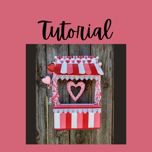 May include: A red, white, and pink kissing booth with a heart-shaped cutout and the word "Tutorial" in black script. The booth has a striped awning and is decorated with hearts and lace trim.