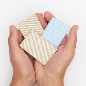 Puede incluir: Tres pequeñas tarjetas rectangulares de papel con diseños estampados se sostienen en manos ahuecadas sobre un fondo blanco. Las tarjetas presentan rayas y un patrón de puntos en varios colores, incluyendo rojo, azul y verde.