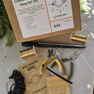 May include: A Crystal Wire Wrap Ring DIY Kit in a cardboard box containing flat nose pliers, wire cutters, round nose pliers, a black ring mandrel, a ring sizer, two spools of gold-colored wire, crystal beads, and an instruction manual.