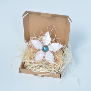 Peut inclure: Une fleur blanche en coquillage et fil de cuivre avec une pierre pr&eacute;cieuse bleue au centre. La fleur est dans une bo&icirc;te en carton marron avec du papier d&eacute;chiquet&eacute;.