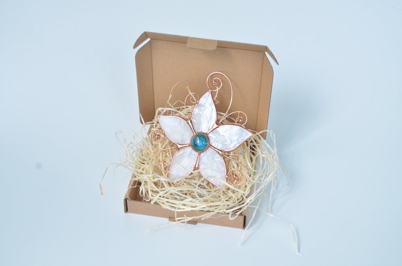 Peut inclure: Une fleur blanche en coquillage et fil de cuivre avec une pierre pr&eacute;cieuse bleue au centre. La fleur est dans une bo&icirc;te en carton marron avec du papier d&eacute;chiquet&eacute;.