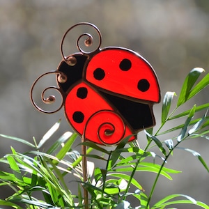 Peut inclure: Un tuteur de jardin en forme de coccinelle rouge et noire, avec une structure en fil de cuivre et des antennes bouclées. La coccinelle a des points noirs et est placée parmi des plantes vertes, sur un fond flou.