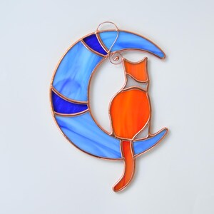 May include: A stained glass cat sitting on a blue crescent moon. The cat is orange with white accents. The moon has blue and purple accents.