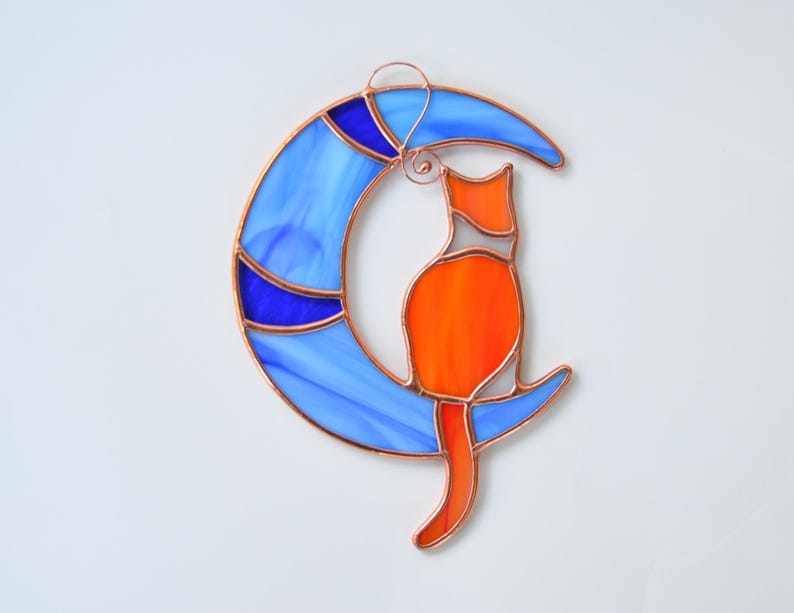 May include: A stained glass cat sitting on a blue crescent moon. The cat is orange with white accents. The moon has blue and purple accents.